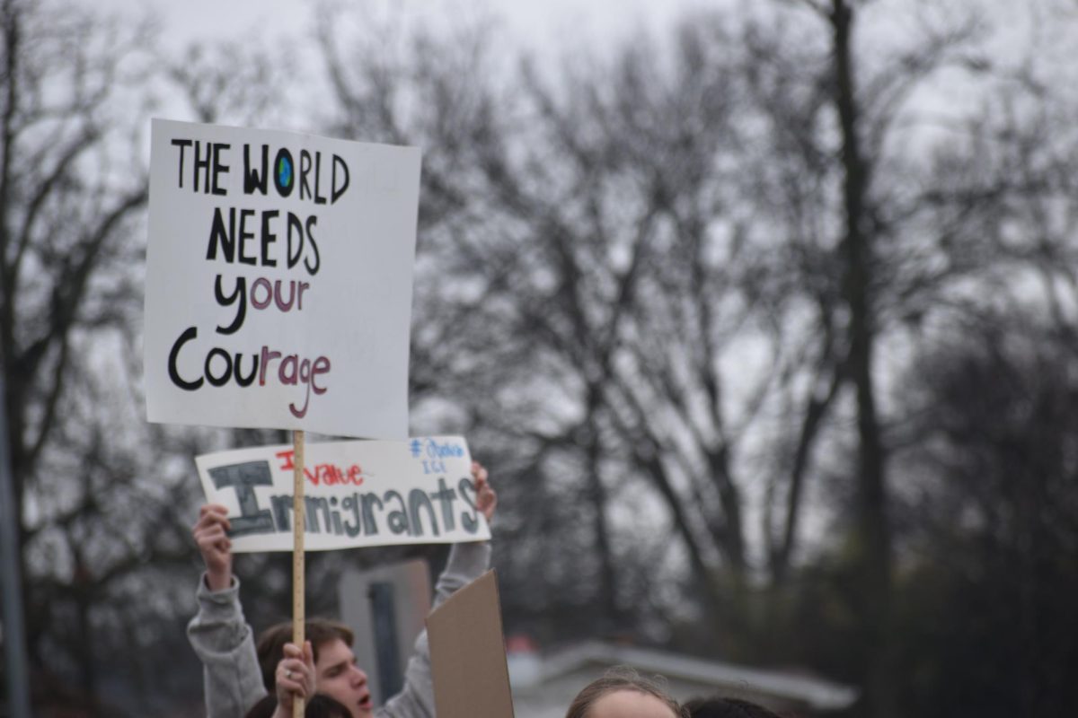 A student holds up a sign saying," The world needs your courage." The words "our rage" in "your courage" are colored differently.